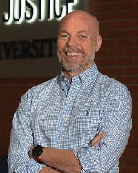 Dr. William Wells poses for a photo. Wearing a button down shirt. Smiling in front of a neon sign glowing Justice.